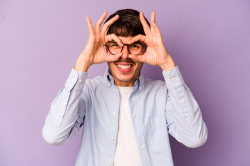 Young caucasian man isolated on purple background showing okay sign over eyes