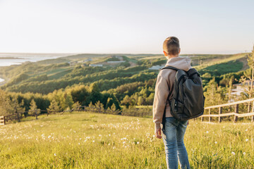 Fototapeta na wymiar Tourist with backpack standing on top of hill in grass field and enjoying beautiful landscape view. Rear view of teenage boy hiker resting in nature. Active lifestyle. Concept of local travel