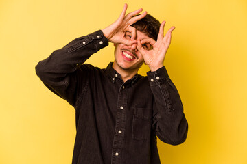 Young caucasian man isolated on yellow background showing okay sign over eyes