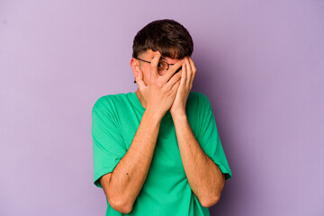Young caucasian man isolated on purple background blink through fingers frightened and nervous.