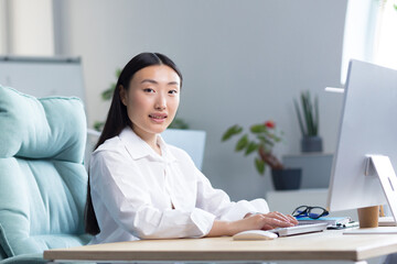 Beautiful Asian woman works at the computer in the office, looks at the camera and smiles, business woman in a white shirt typing on the keyboard.