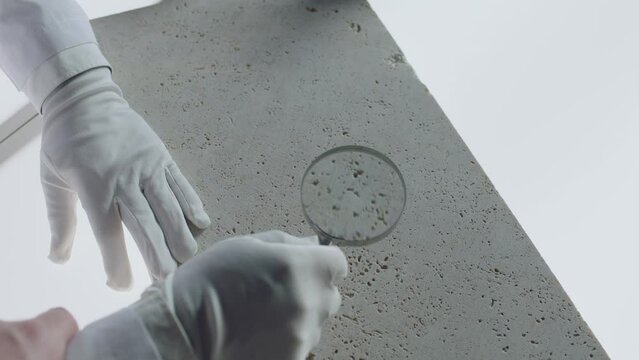 Construction laboratory testing the quality density of natural travertine stone . Worker of laboratory explore stone by loupe . Close up view of hands with gloves . Shot on ARRI Alexa cinema camera