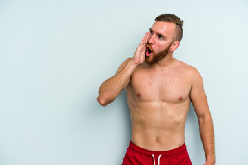 Young caucasian man wearing a swimsuit isolated on blue background is saying a secret hot braking news and looking aside