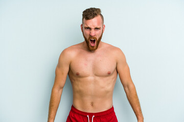 Young caucasian man wearing a swimsuit isolated on blue background screaming very angry and aggressive.