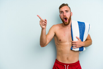 Young caucasian man going to the beach holding a towel isolated on blue background pointing to the side