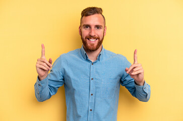 Young caucasian man isolated on yellow background indicates with both fore fingers up showing a...