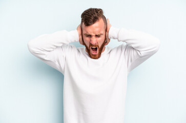 Young caucasian man isolated on blue background covering ears with hands trying not to hear too loud sound.