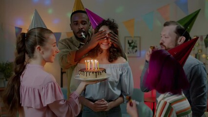 Young woman celebrating birthday blowing candles on cake while friends in party hats blowing horns and clapping hands. Youth and holiday celebration concept.
