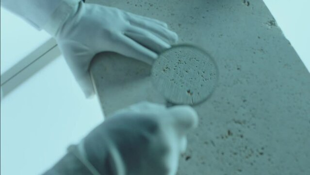 Construction laboratory testing the quality density of natural travertine stone . Worker of laboratory explore stone by loupe . Close up view of hands with gloves . Shot on ARRI Alexa cinema camera