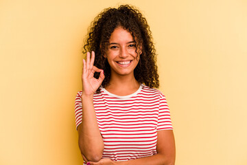 Young Brazilian woman isolated on yellow background winks an eye and holds an okay gesture with hand.