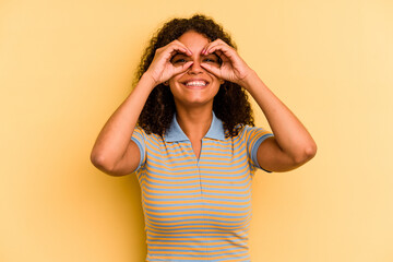 Young Brazilian woman isolated on yellow background showing okay sign over eyes