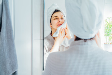 Woman in bathrobe with a towel on head looking in the mirror and drying her face with a towel in...