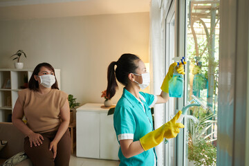 Young woman from cleaning service washing windows in the room with owner watching for process