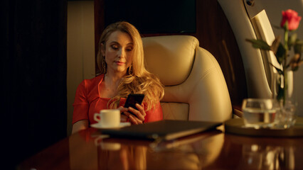 Blonde lady messaging mobile phone in private airplane in golden sunlight.