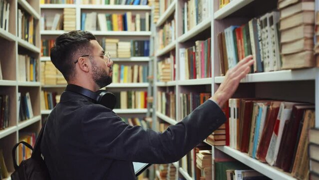 Young man with beard wearing eyeglasses and having headphones on neck standing by bookcase and choosing books, checking list of literature on tablet, student in library. Concept of education