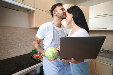Caring husband planting kiss on forehead of wife with laptop