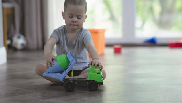 little boy child sitting on wooden floor in living room playing plastic cement mixer toy car. construction cite concept baby kid imagine pretending being driver worker. leisure activity children