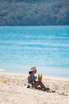 Vertical image of a woman on the beach wearing a hat and sunglasses, sipping a delicious drink on a summer day. 