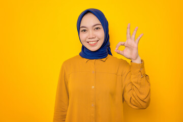 Cheerful young Asian Muslim woman dressed in orange shirt showing okay gesture isolated on yellow background