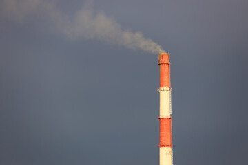 High red and white chimney with smoke against a dark blue stormy sky. Atmospheric pollution with industrial emissions, environmental degradation and greenhouse gases concept