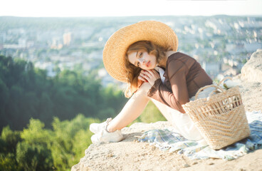 Portrait of a blonde girl in a straw hat posing with her arms around her knees against the...