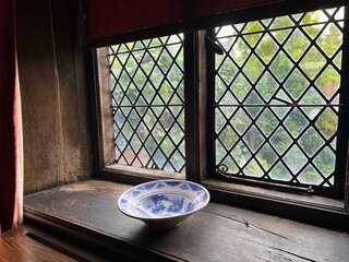 Close up of medieval Elizabethan leaded glass window diamond shape thick hand blown historic glass  light  shadows on ancient historic wood window cill with antique vintage china bowl with tree nuts