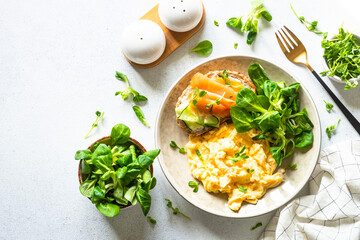 Scrambled eggs, sandwich with cream cheese and salmon and salad leaves at white stone table. Top...