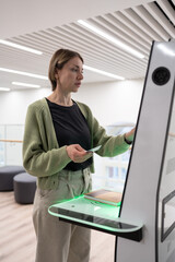 Middle-aged scandinavian woman using library self-service machine for borrow or return book, female...