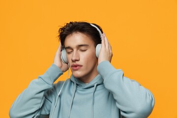 a happy man in a blue hoodie stands on an orange background listening to music with headphones holding them with his hands closing his eyes with pleasure - Powered by Adobe