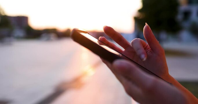 Hands of young woman using smartphone at sunset, close-up