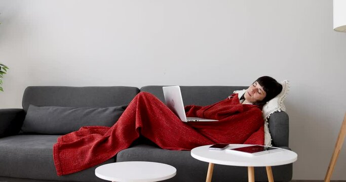 Tired woman using laptop lying on couch wrapped in blanket