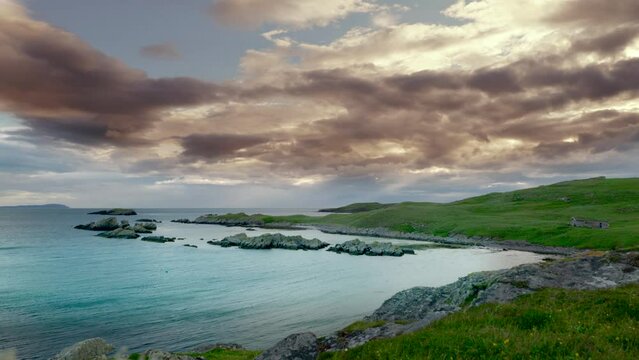 Breathtaking timelapse of fast moving clouds during an orange and gold sunset over the lush green grass of Handa Island in Scotland, with turquoise waves lapping against the jagged rocky shoreline