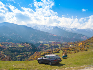 Man and a woman are sitting on chairs near the car and enjoying the amazing beautiful autumn mountain landscape of Svaneti, view from the drone. Concept of a tourist lifestyle, traveling by car.