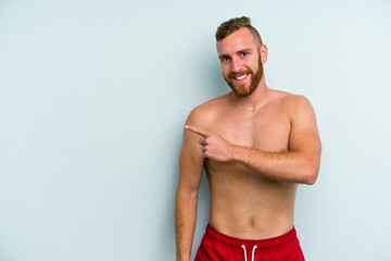 Young caucasian man wearing a swimsuit isolated on blue background smiling and pointing aside, showing something at blank space.