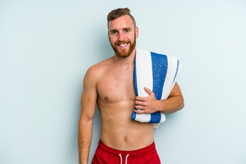 Young caucasian man going to the beach holding a towel isolated on blue background happy, smiling and cheerful.