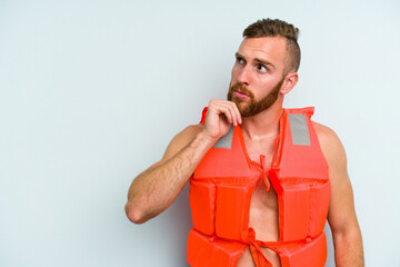 Young caucasian man wearing life jacket isolated on blue background looking sideways with doubtful...