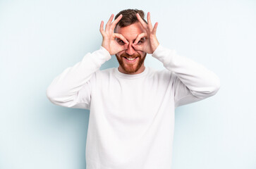 Young caucasian man isolated on blue background showing okay sign over eyes
