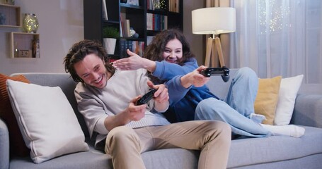 Happy young couple holding wireless joysticks and playing game at home during evening time. Caucasian man and woman enjoying leisure activity while sitting on sofa in front of TV. - Powered by Adobe