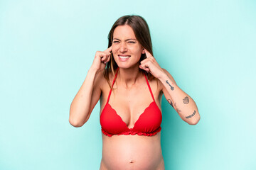 Young caucasian pregnant woman wearing bikini isolated on blue background covering ears with hands.