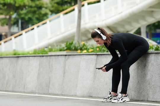 Young sportive girl in wireless headhones tracking her workouts on smartphone while sitting outdoors and resting after running