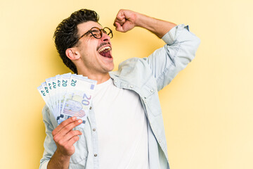 Young caucasian man holding banknotes isolated on yellow background raising fist after a victory,...