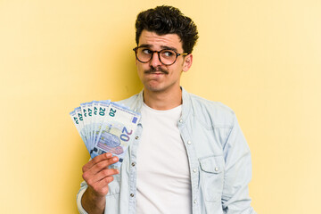 Young caucasian man holding banknotes isolated on yellow background confused, feels doubtful and unsure. - obrazy, fototapety, plakaty