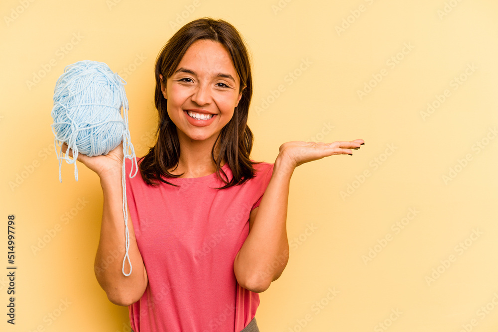 Wall mural Young hispanic woman holding a ball of wool isolated on yellow background showing a copy space on a palm and holding another hand on waist.