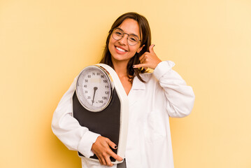 Young nutritionist woman holding a weighing machine isolated on yellow background showing a mobile...