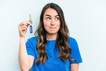Young caucasian woman holding home keys isolated on blue background confused, feels doubtful and unsure.