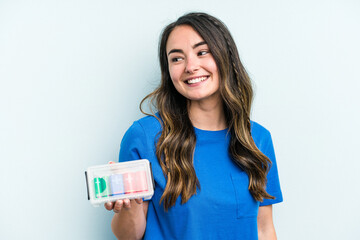 Young caucasian woman holding batteries isolated on blue background looks aside smiling, cheerful and pleasant.