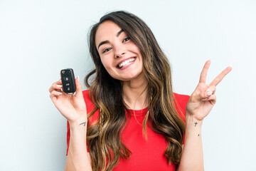 Fototapeta premium Young caucasian woman holding car keys isolated on blue background joyful and carefree showing a peace symbol with fingers.