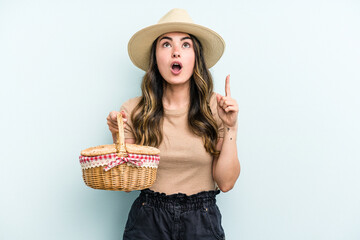 Young caucasian woman holding a picnic basket isolated on blue background pointing upside with opened mouth.