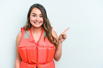 Young caucasian woman wearing life jacket isolated on blue background smiling and pointing aside,...