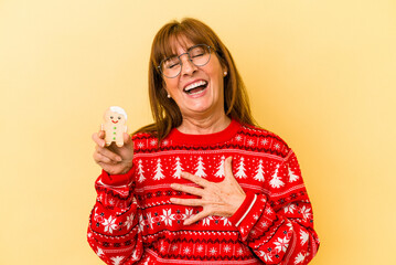 Middle age caucasian woman holding a Christmas cookies isolated on yellow background .laughs out loudly keeping hand on chest.